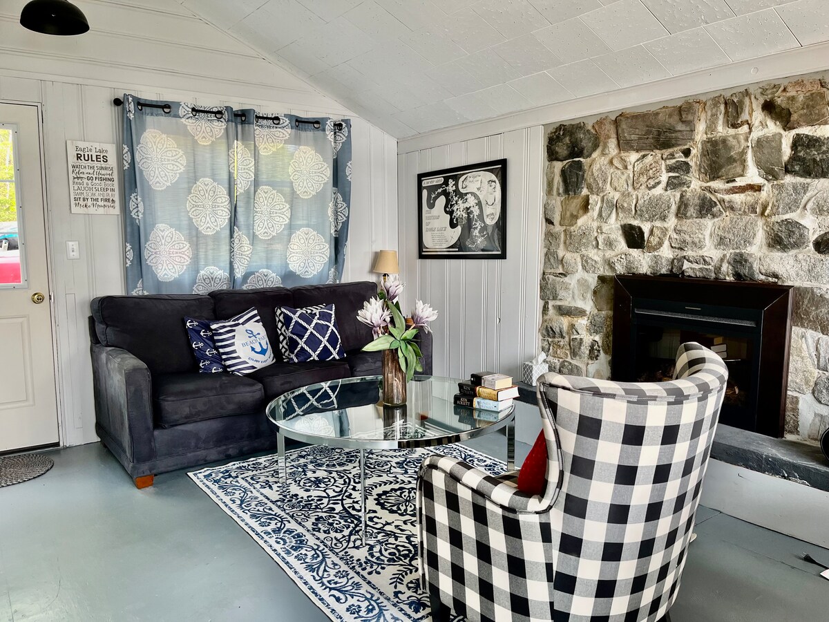A cozy living area features a dark sofa adorned with decorative pillows. A round glass coffee table sits atop a patterned rug, complemented by a checkered armchair. A rustic stone fireplace creates a focal point, while curtains add a touch of warmth to the space.