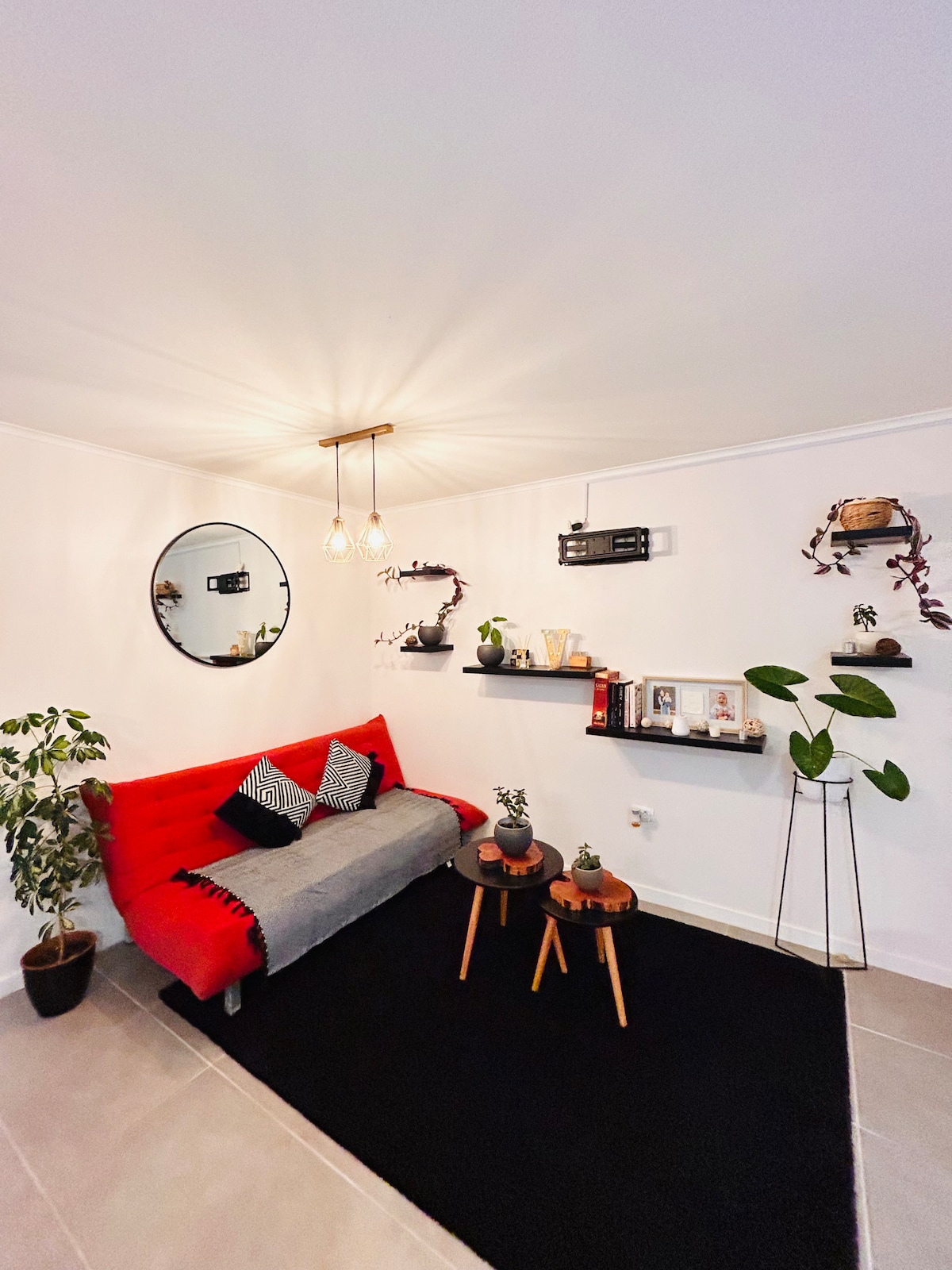 A inviting living area is furnished with a red sofa adorned with cushions, complemented by small wooden tables. Shelves display decorative items and plants, while a round mirror enhances the space. Soft lighting is provided by hanging fixtures, adding warmth to the minimalist decor.