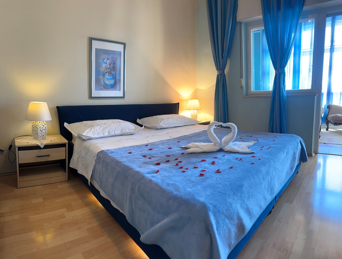 A spacious bed is centered in the room, adorned with a light blue blanket and decorative towels shaped like swans. Soft light is provided by bedside lamps, and bright blue curtains frame the window, allowing natural light to illuminate the space. A piece of framed artwork is hung on the wall.