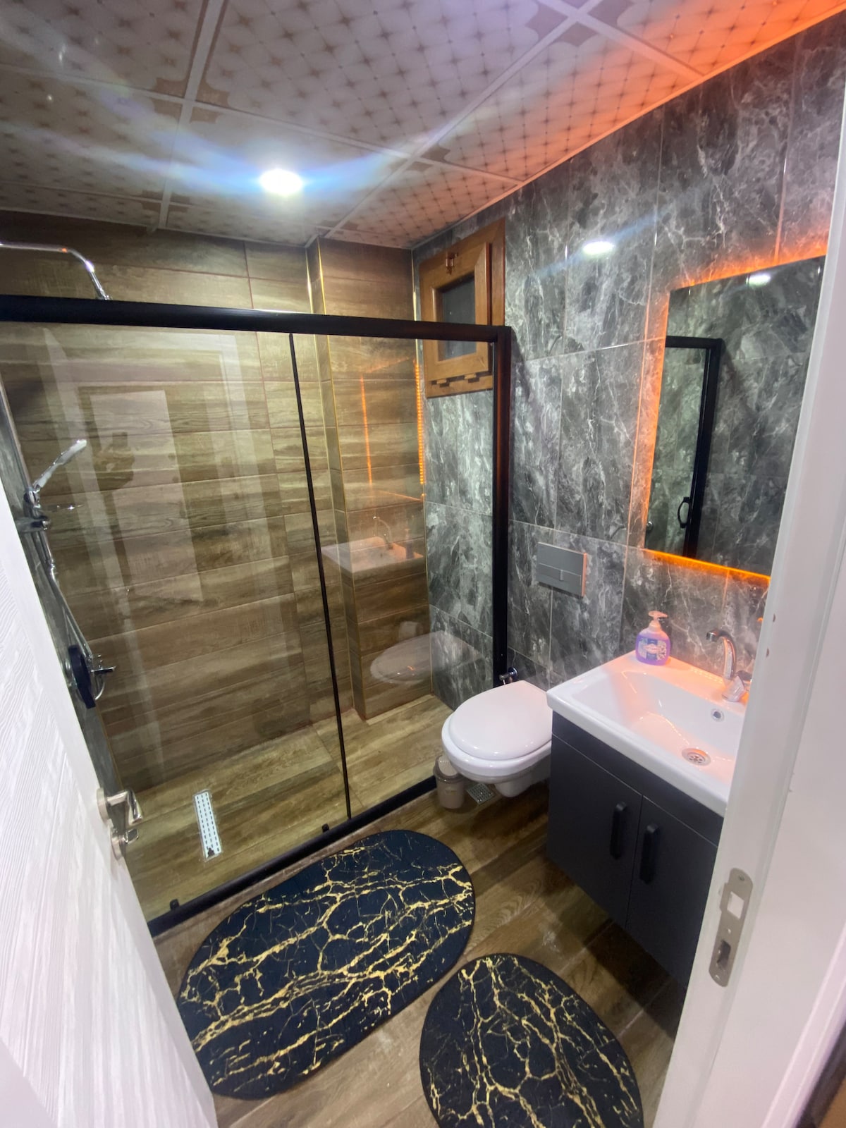 A modern bathroom features a glass shower enclosure and stylish grey marble tiles. A white sink is positioned beside a wall-mounted mirror, and decorative mats with a marble pattern are placed on the floor. Soft lighting highlights the contemporary design.