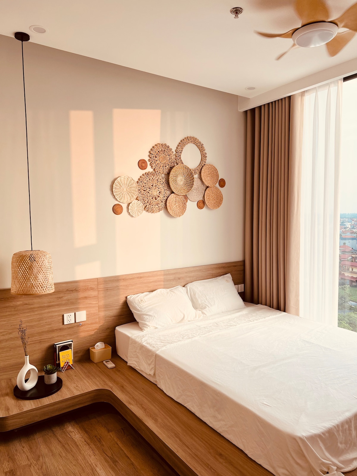 A serene bedroom is illuminated by natural light filtering through large windows. A spacious bed with a white duvet is positioned against a wooden wall. Decorative wall art made of woven materials adds texture to the space. A bedside table holds a white vase and a small lamp.
