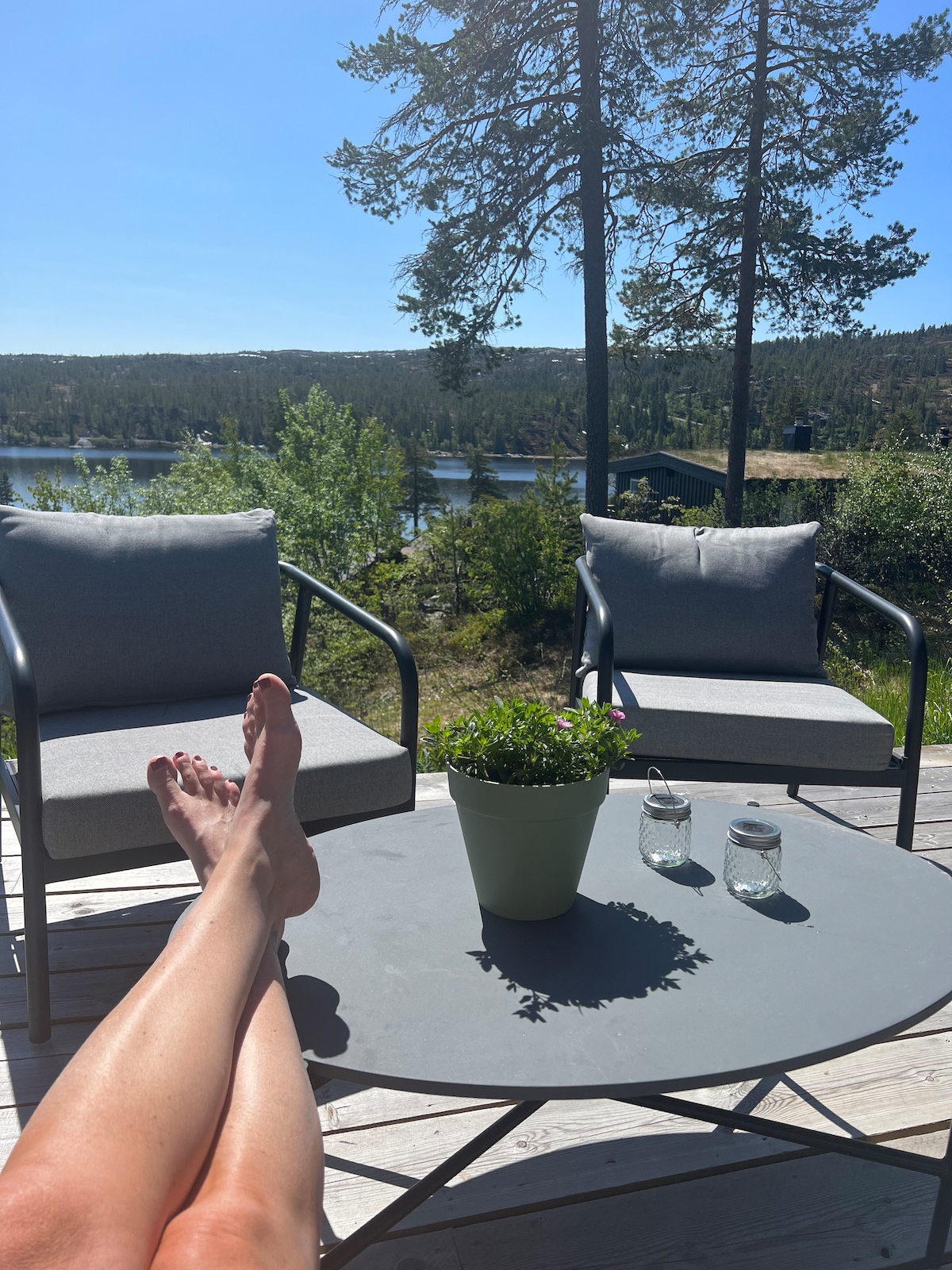 An outdoor space is shown, featuring two comfortable chairs positioned beside a round table. A potted plant sits centrally, accompanied by glass jars. In the background, lush greenery and a serene body of water create a tranquil setting under clear blue skies.
