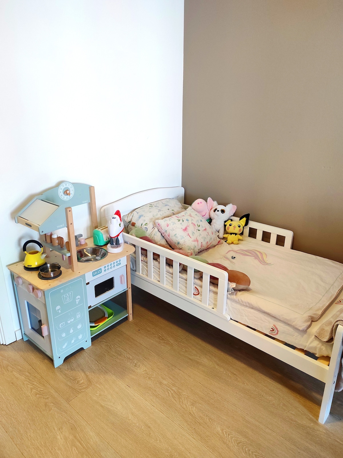 A children's sleeping area features a small bed with light bedding and plush toys. Adjacent to the bed is a wooden play kitchen, equipped with assorted cooking utensils and appliances, enhancing imaginative play opportunities.
