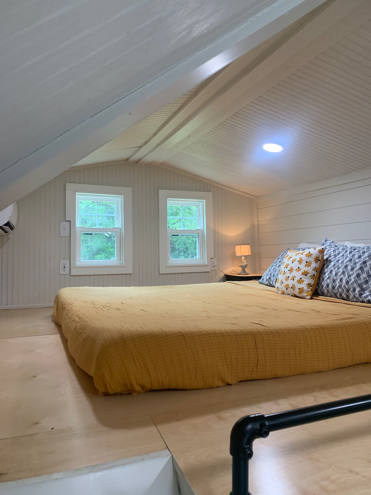 A cozy sleeping loft is showcased, featuring a spacious bed with a mustard yellow coverlet and decorative pillows. Two windows on one side provide natural light, while a small lamp adds a gentle glow. The ceiling is lined with wooden paneling, enhancing the airy feel.