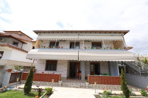 A two-story house with a covered balcony is presented, featuring light-colored walls and terracotta accents. Potted plants adorn the balcony railing. A spacious entrance is visible, complemented by well-maintained garden areas including neatly trimmed shrubs and flower beds.