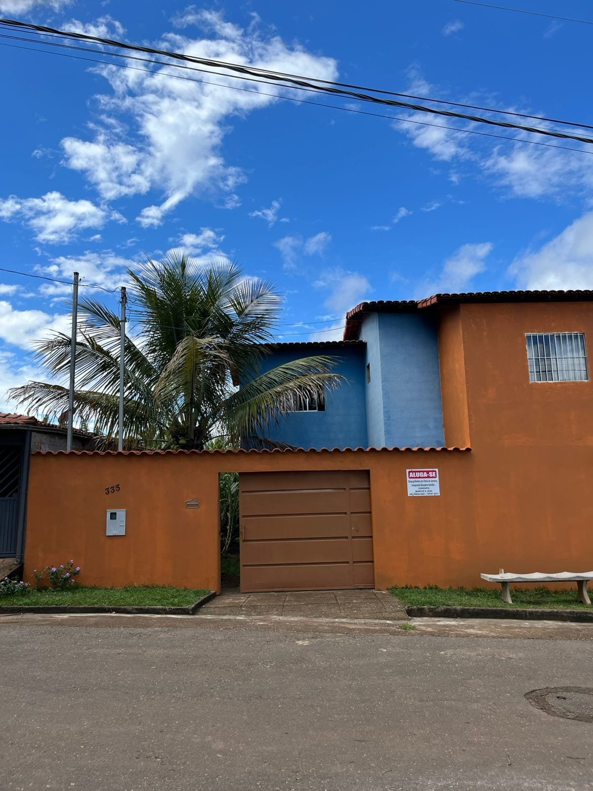 The exterior of a vibrant orange house is presented, featuring a blue section at the top. Tall palm trees add greenery to the surroundings, while the clear blue sky enhances the overall brightness. A metal gate is positioned in front, with a seating area nearby.