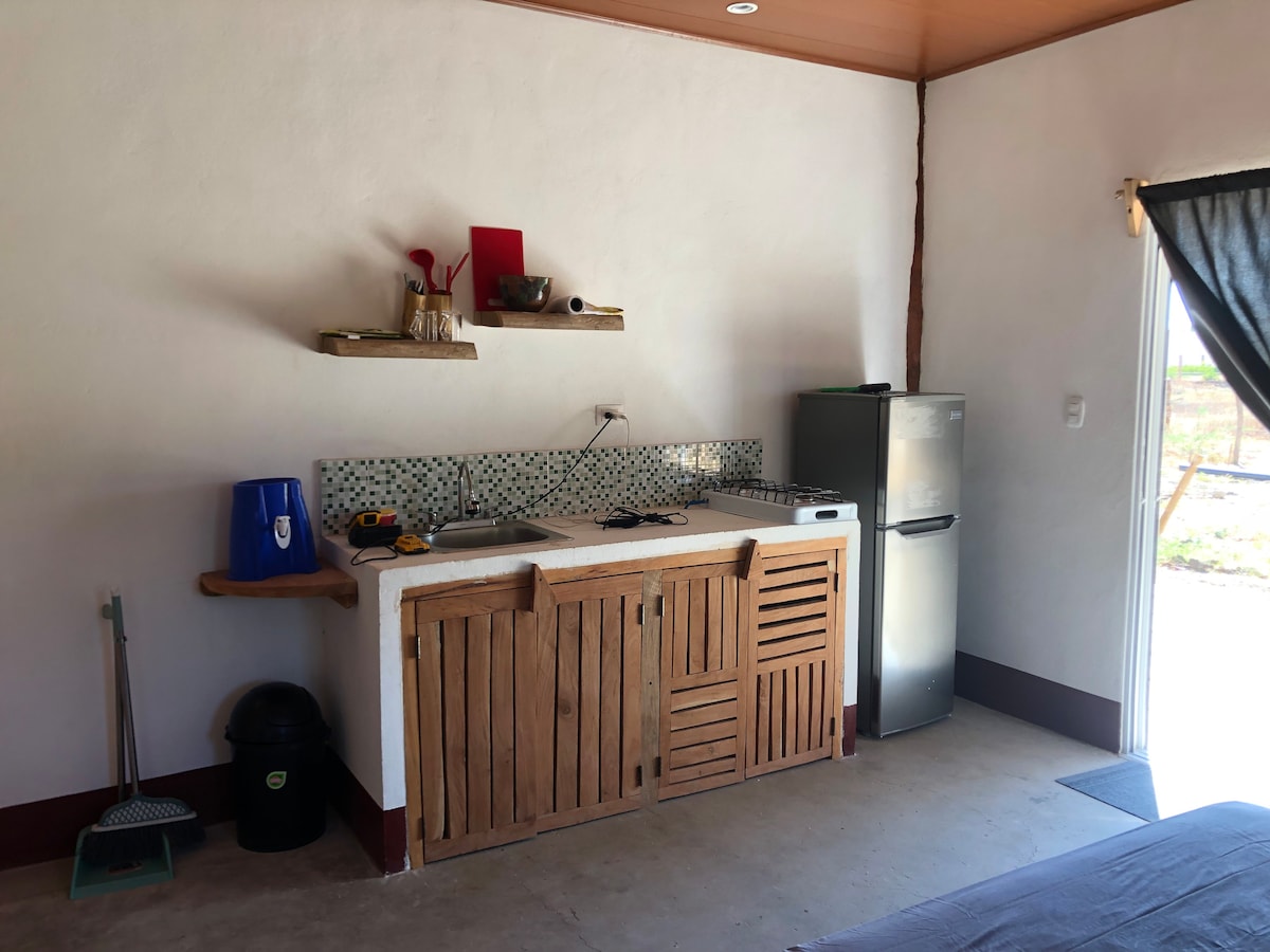 A functional kitchen area is presented with a wooden counter featuring open shelving above. A small refrigerator and a sink are included, complemented by a tiled backsplash. Cooking utensils and a red cutting board are visible, along with cleaning supplies placed in the corner.