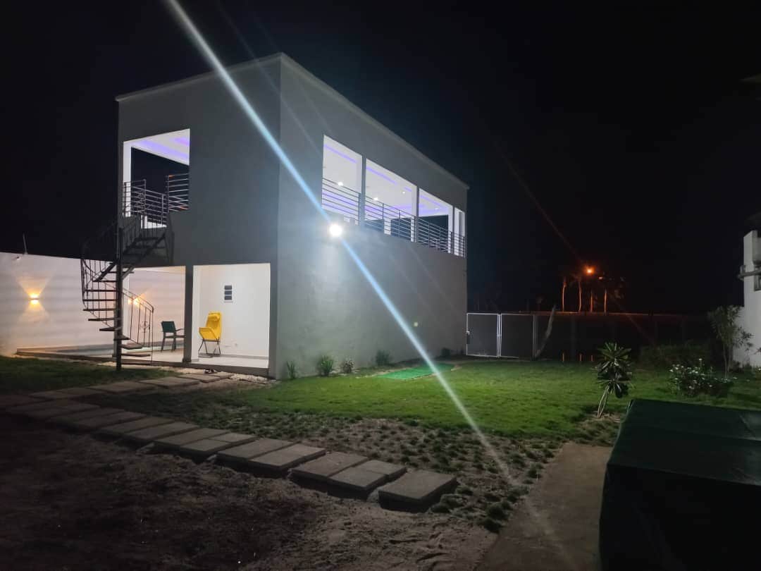 A modern two-story building is illuminated by exterior lights, highlighting the spacious yard and well-maintained grass. A staircase leads to a wraparound balcony, while a few chairs are positioned nearby. Soft ambient lighting enhances the evening atmosphere.