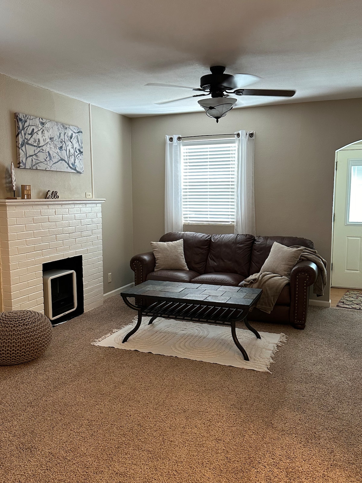 The living area features a comfortable brown leather sofa paired with a textured coffee table. A cozy area rug lies on the carpeted floor, while a white-brick fireplace offers a warm focal point. Natural light enters through a window adorned with light curtains.