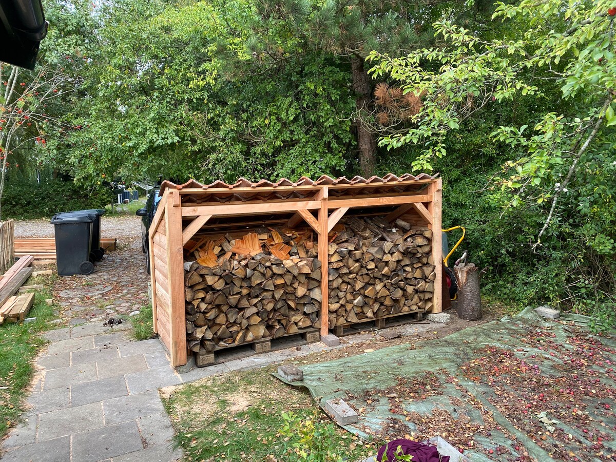 A wood shed is neatly organized, holding stacked firewood beneath a sloped roof. Nearby, a tarpaulin covers a patch of ground, while trees and shrubbery create a green backdrop. A compost bin can be seen to the right, contributing to the natural ambiance of the outdoor space.