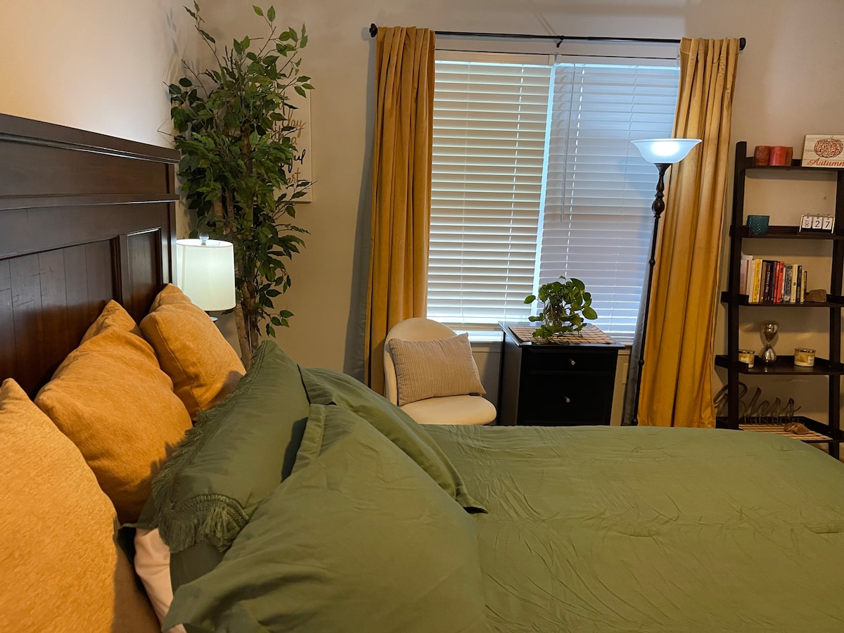 A cozy bedroom includes a spacious bed dressed in calming green linens and several yellow accent pillows. Natural light streams through a window adorned with light-filtering blinds, while a plant adds a touch of greenery. A small armchair sits beside a nightstand.