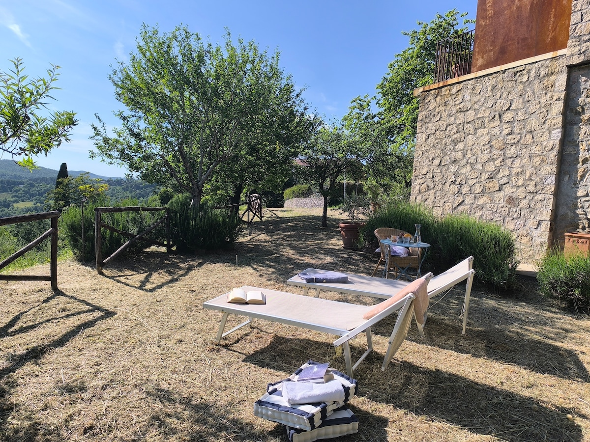 A tranquil outdoor area is presented, featuring two lounge chairs positioned on a grassy surface. Surrounding greenery includes shrubs and small trees, while a distant view of rolling hills is visible. A small table sits nearby, adding to the inviting space.