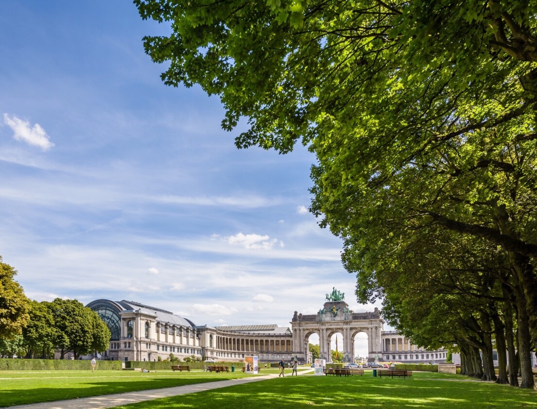 A green park area is framed by tall trees on either side, leading toward an impressive archway. The clear blue sky is dotted with clouds, and the expansive grass area invites relaxation and outdoor activities. Architectural elements of the nearby buildings are visible in the background.