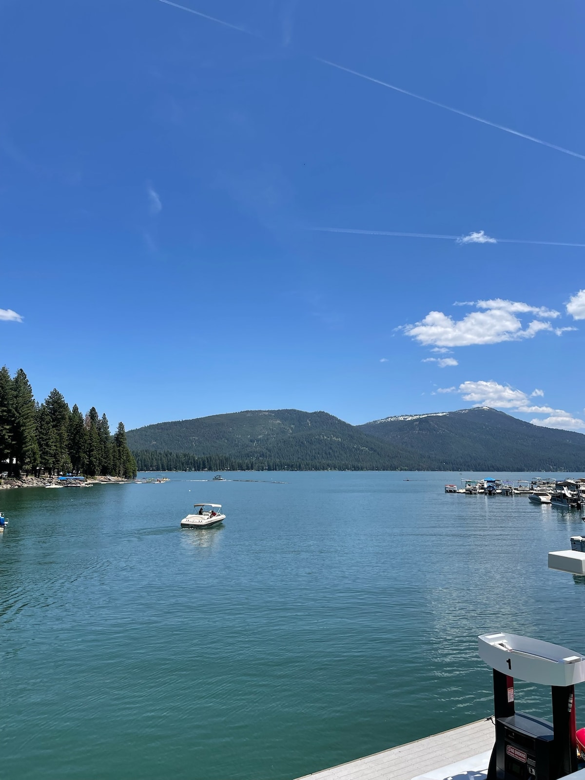 A serene lake scene is captured, showcasing calm waters reflecting a clear blue sky. Lush green trees line the shores, while distant mountains provide a picturesque backdrop. Several boats are visible on the water, indicating a tranquil day for recreation.