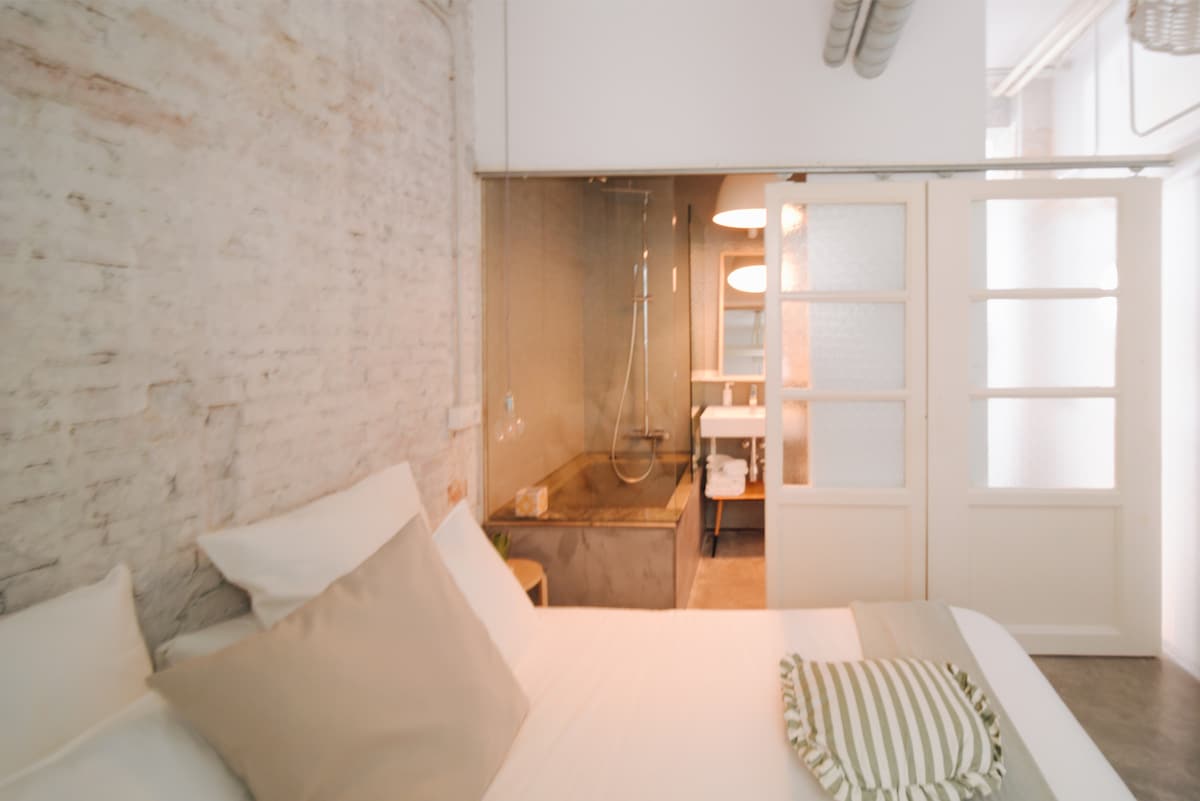 A serene bedroom features a spacious bed with soft linens and accent pillows. The open layout reveals a modern bathroom area in the background, highlighted by a glass partition and stylish fixtures. Exposed brick walls add character to the calming space.