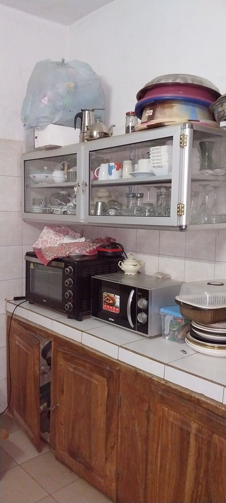 A compact kitchen area features wooden cabinets with glass-front doors showcasing various dishes and glassware. A microwave and an oven are positioned on the countertop, alongside a collection of bowls and utensils. Light-colored tiles create a clean, functional space.