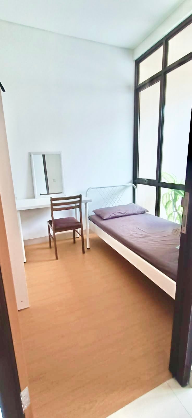 A bright room features a spacious bed with a simple purple bedding set. A wooden chair is positioned nearby, alongside a minimalist desk with a mirror. Large windows allow natural light to fill the space, complemented by a small plant in the corner.