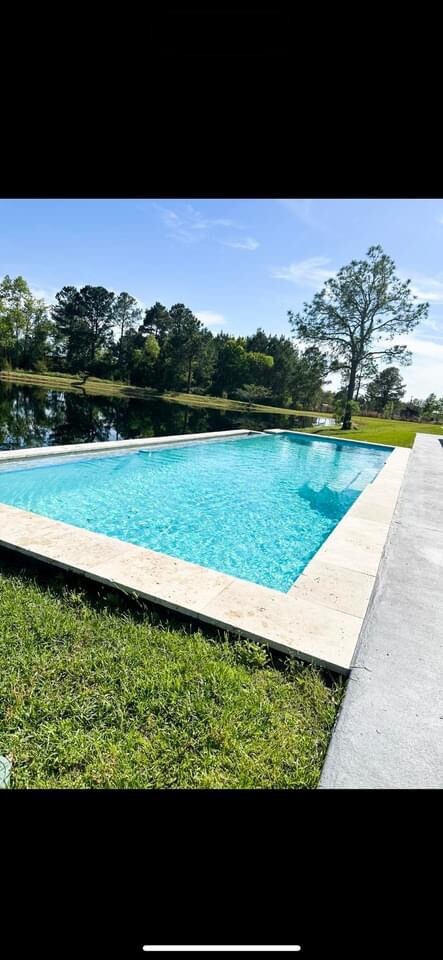 A tranquil pool is surrounded by a clean, light-colored stone deck. The clear blue water reflects sunlight and features a lush green landscape in the background, with trees and grass providing a serene setting.