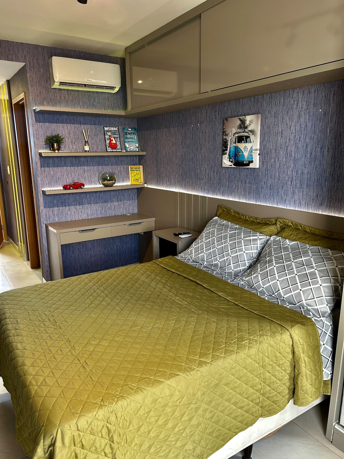 A well-appointed bedroom is arranged with a spacious bed covered in a soft gold quilt. Grey patterned pillows provide contrast against the quilt. A modern shelf displays decorative books and a small plant. Natural light filters through a nearby window, and an air conditioning unit is mounted above.