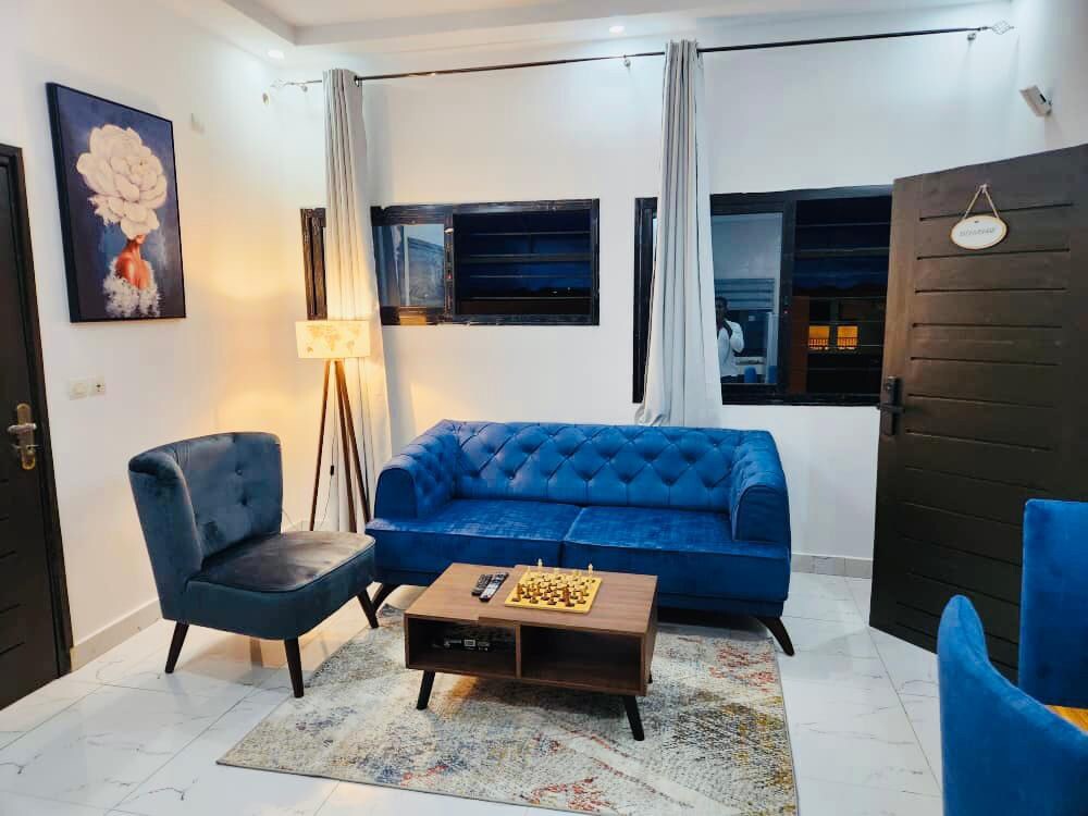 A modern living area showcases a deep blue tufted sofa and a plush armchair in a coordinating shade. A wooden coffee table is centered on a patterned rug. Soft lighting from a floor lamp illuminates the space, while large windows create an airy feel.