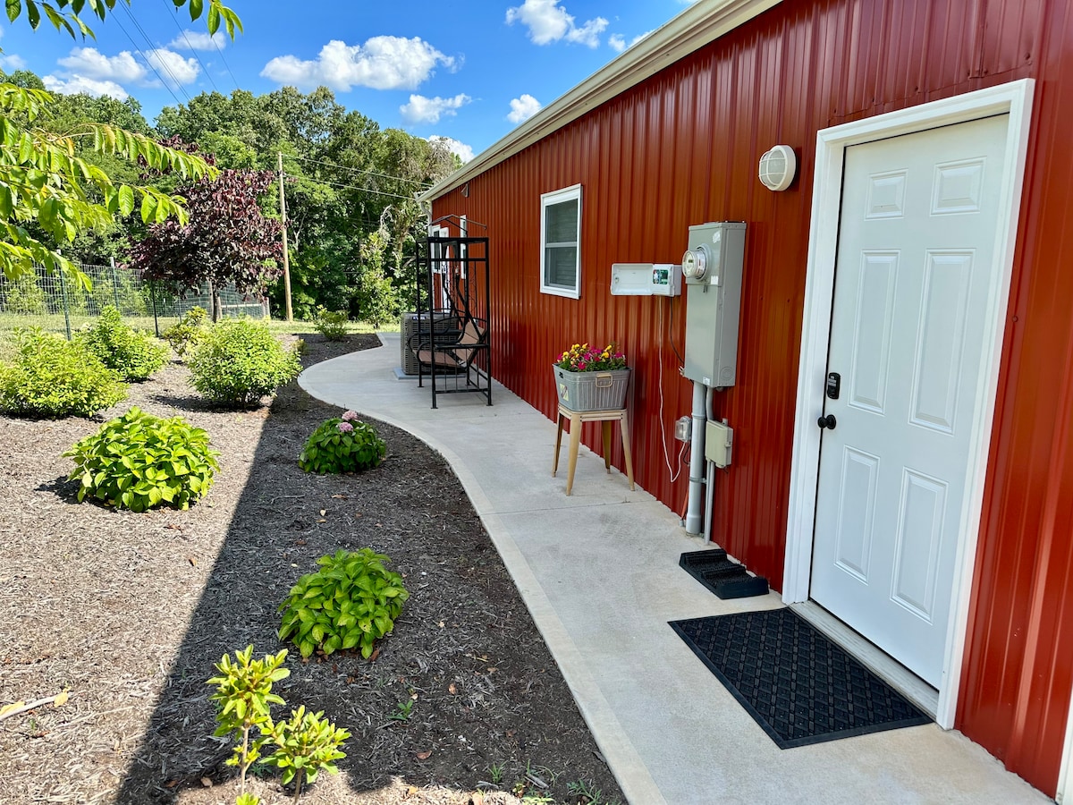 A view of the guesthouse exterior features a red metal wall and a welcoming entrance. A concrete pathway leads to the door, bordered by mulched garden beds with greenery. A seating area with black chairs and a small table is visible nearby, inviting relaxation outdoors.