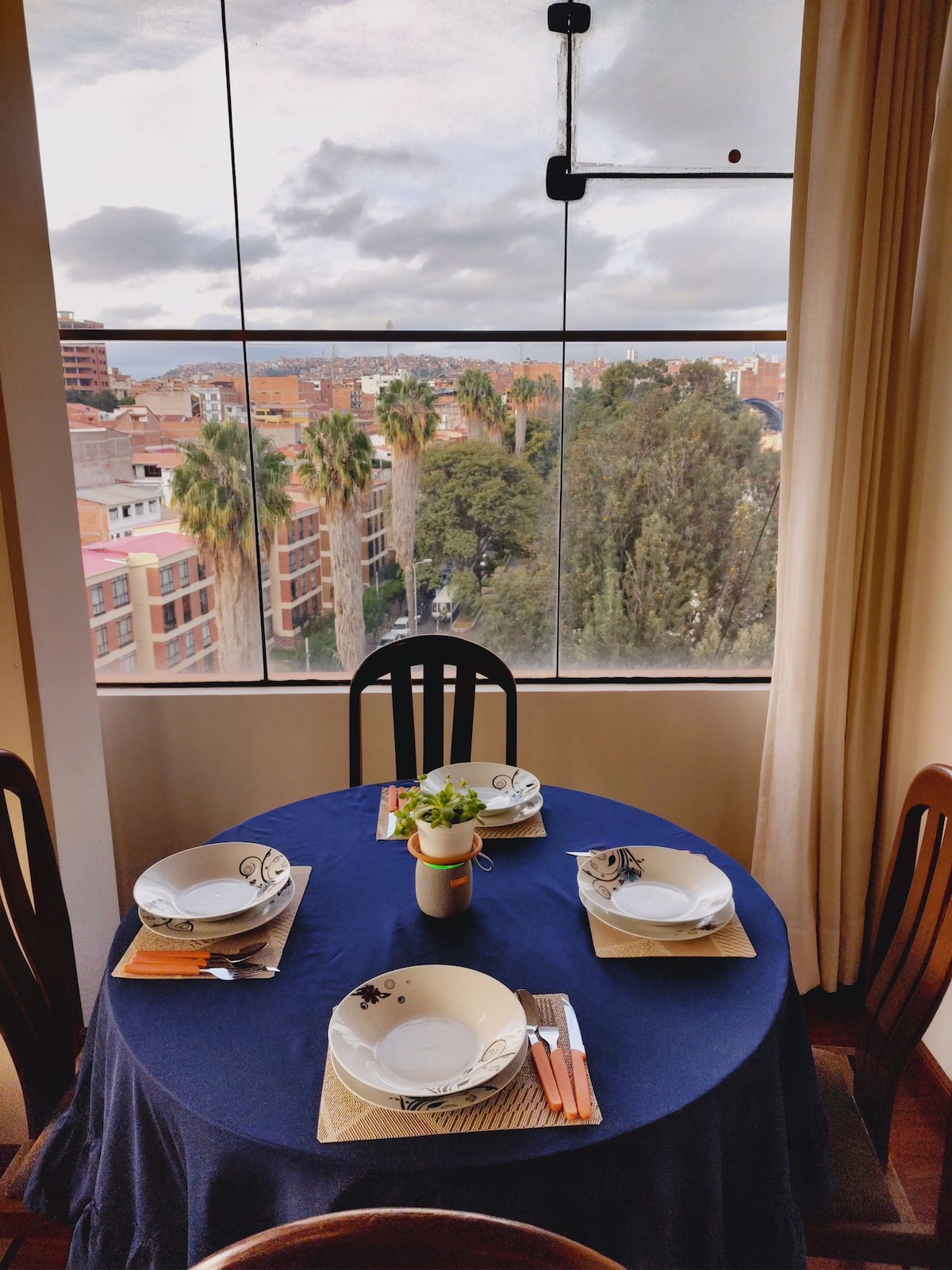 A dining table is set with four plates, cutlery, and a small plant centerpiece. The table is covered with a dark blue tablecloth. Large windows provide a view of palm trees and the city skyline, allowing natural light to fill the space.