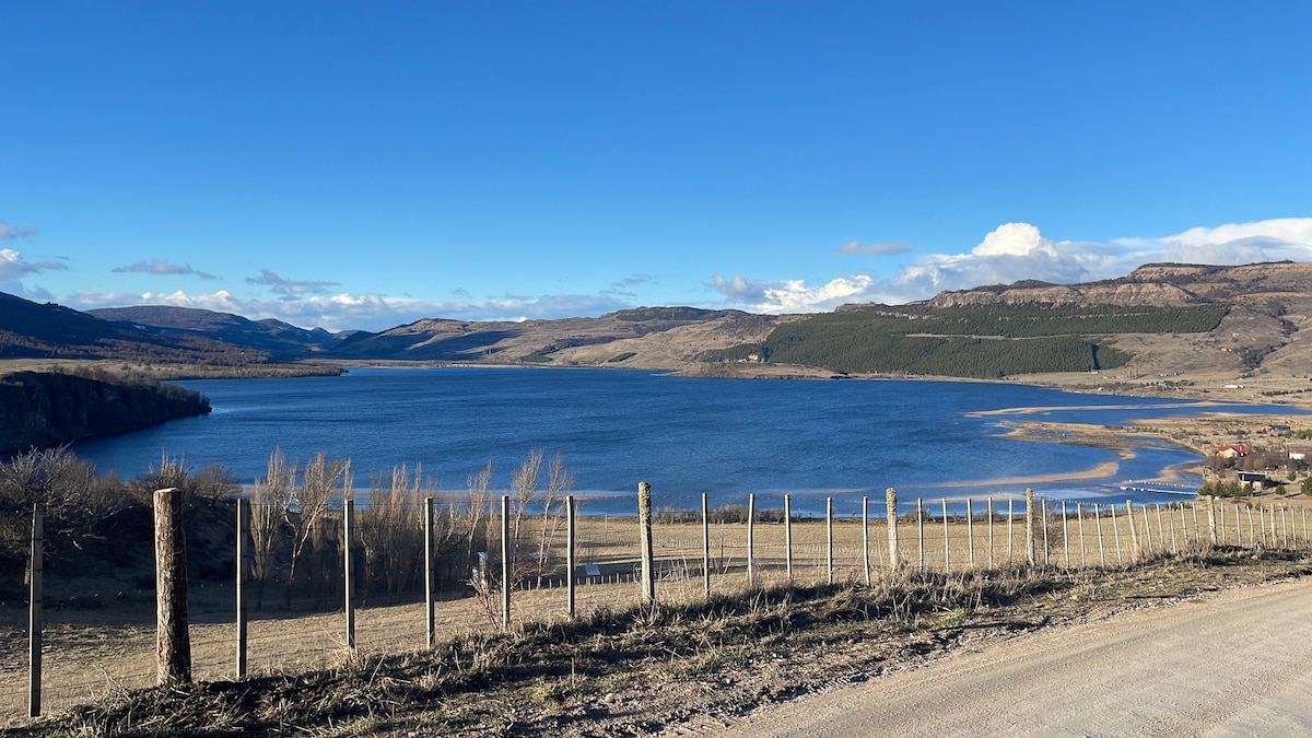 A scenic view of a calm lake is depicted, surrounded by rolling hills and mountains under a clear blue sky. The landscape includes a row of wooden fence posts along the road, leading towards the tranquil water's edge.