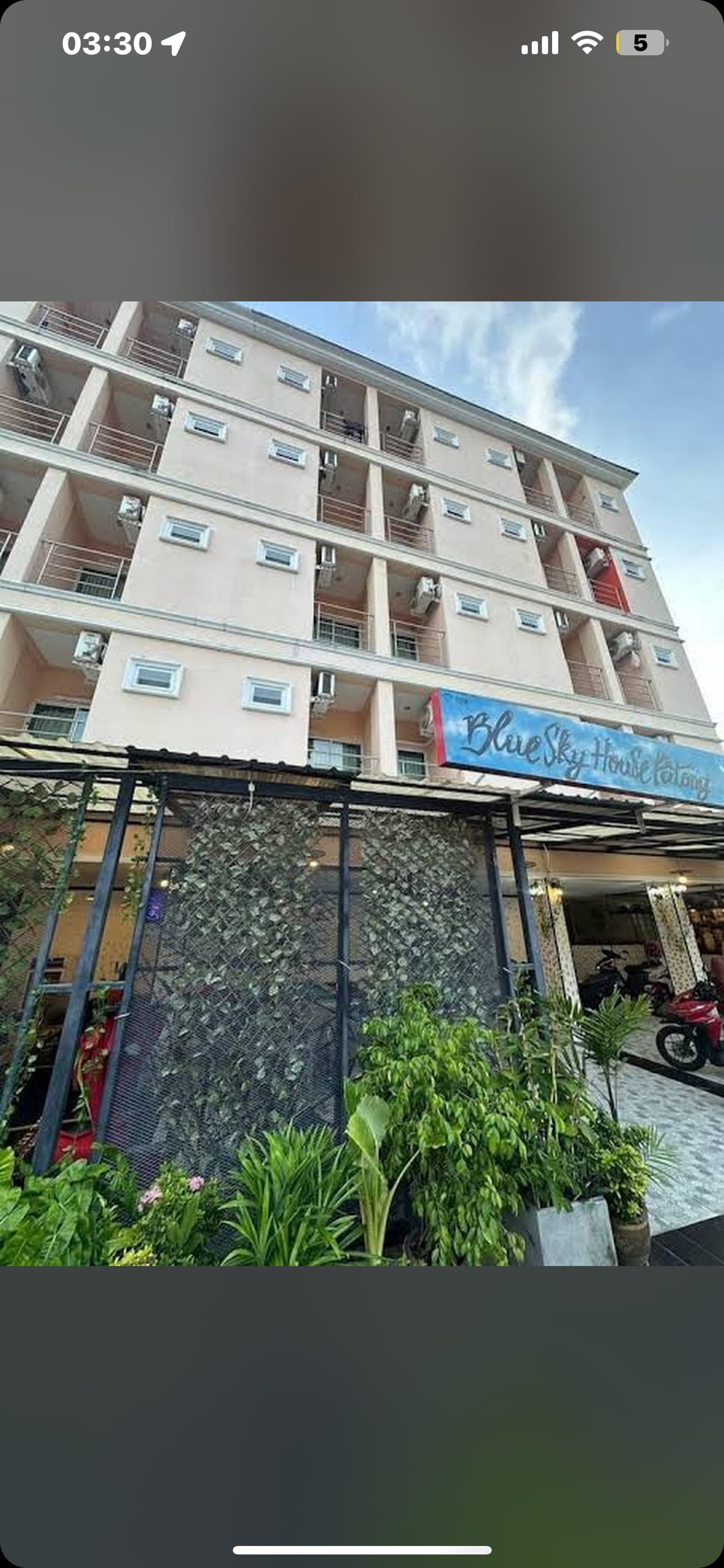 A multi-story building is shown, featuring a light pink facade with multiple windows. The entrance is framed by a decorative green wall, alongside potted plants. A sign reading 'Blue Sky Hotel' is prominently displayed above the entrance, welcoming visitors.