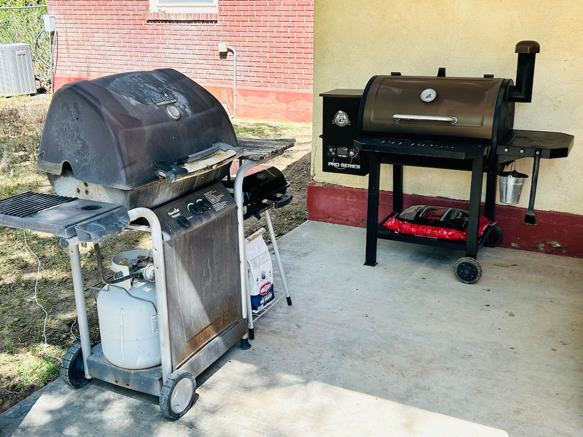 Two grills are positioned on a concrete surface under a shaded area. One is a traditional gas grill with a propane tank, while the other is a smoker grill, both designed for outdoor cooking. Surrounding greenery is visible in the background.