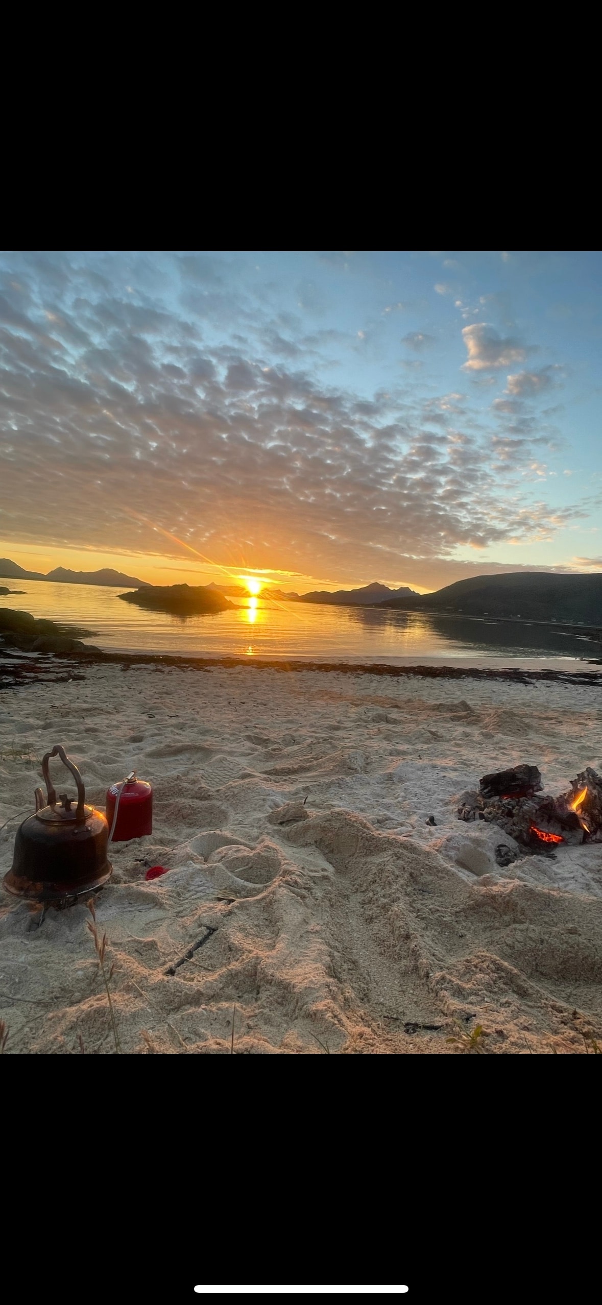 A serene beach scene showcases soft, white sand leading to a tranquil body of water. The sun sets behind distant mountains, casting warm tones across the sky and reflecting on the water's surface. A small fire pit is visible, surrounded by scattered stones.