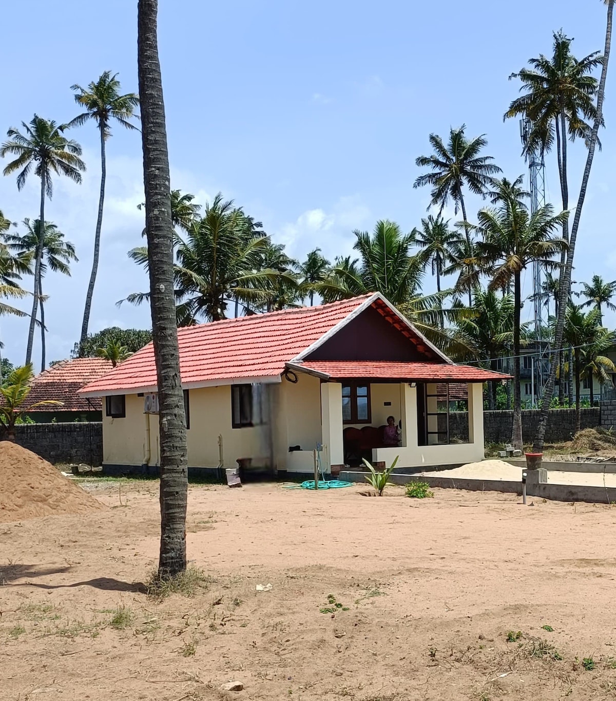 A charming beach house is surrounded by palm trees, featuring a red-tiled roof and large windows. The property is set in a sandy area with landscaping elements visible. The structure provides a sense of openness and tranquility, inviting guests to enjoy the natural environment.