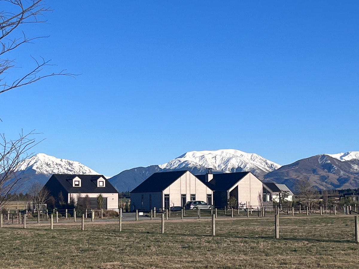 A view of the property showcases two buildings in a serene landscape, framed by the snow-capped mountains in the background. The open field highlights the spaciousness of the surroundings, with a clear blue sky overhead and a rustic fence dividing the area.