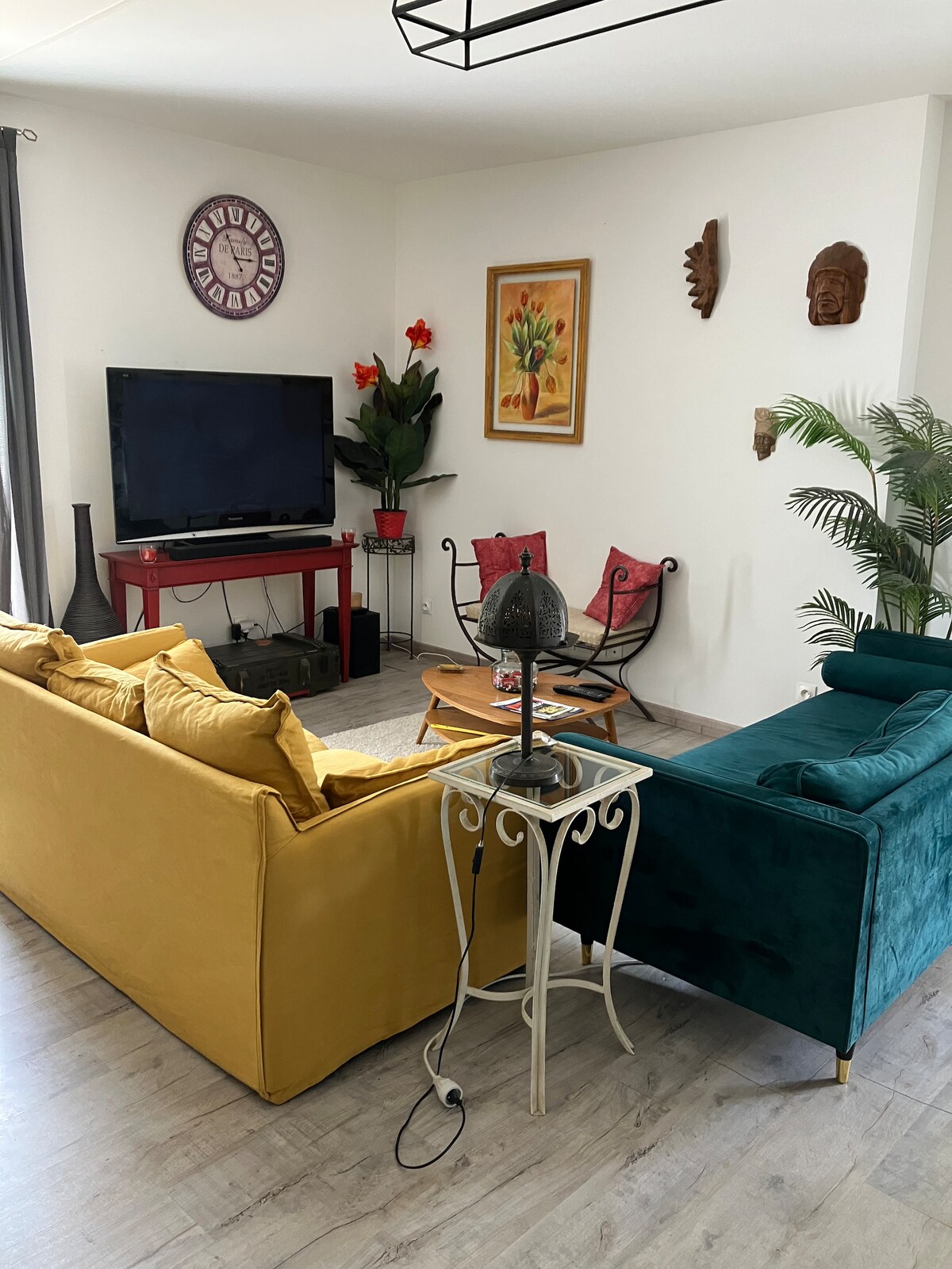 A cozy living area is presented, featuring a yellow modular sofa and a teal armchair, creating a vibrant contrast. A wall-mounted television is visible above a red media console, while a decorative table holds a plant and magazines. Artwork and plants add character to the space.