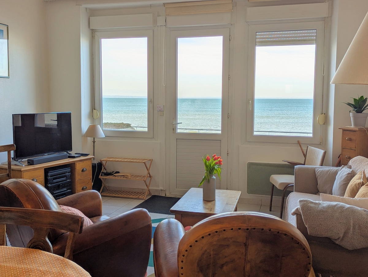 A living area is presented with multiple windows, offering a view of the sea. Comfortable seating arrangements include wooden chairs and a soft sofa. A television is placed on a wooden unit, and a vase of flowers sits centered on a coffee table.