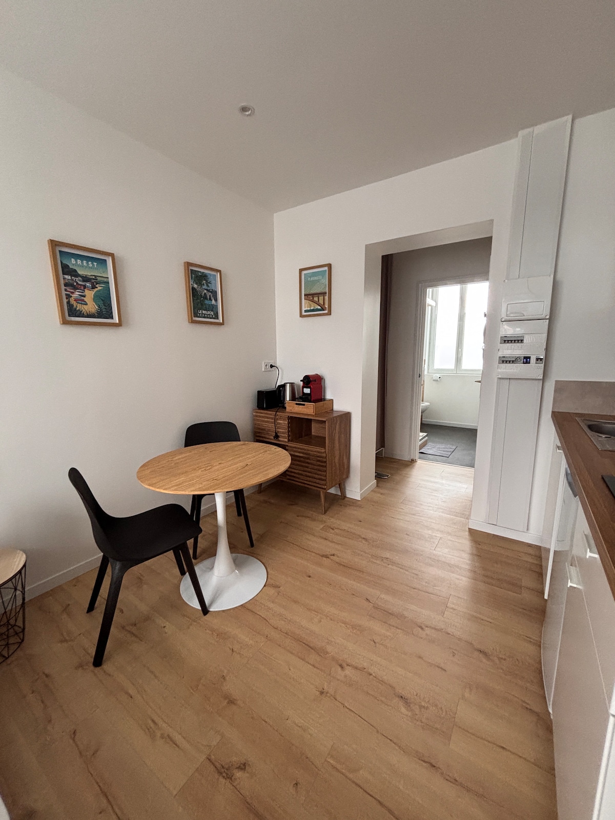 A small kitchen area is presented, featuring a round wooden table with two black chairs. A console with a coffee maker and a water kettle is positioned against the wall. Two framed artworks are displayed on the adjacent wall, complementing the light wooden flooring.