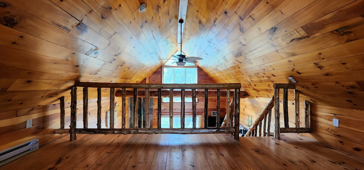 The cozy loft area features a wooden railing overlooking the open-plan living space below. Natural light floods in through large windows, highlighting the warm wooden ceiling and floor. A ceiling fan is installed for comfort in the rustic retreat.