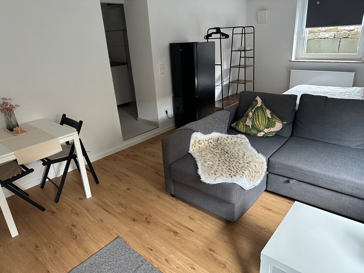 A cozy living area features a gray sectional sofa draped with a soft throw. A small dining table with black folding chairs is positioned nearby. The space showcases light wooden flooring and bright walls, with a kitchen area visible in the background.