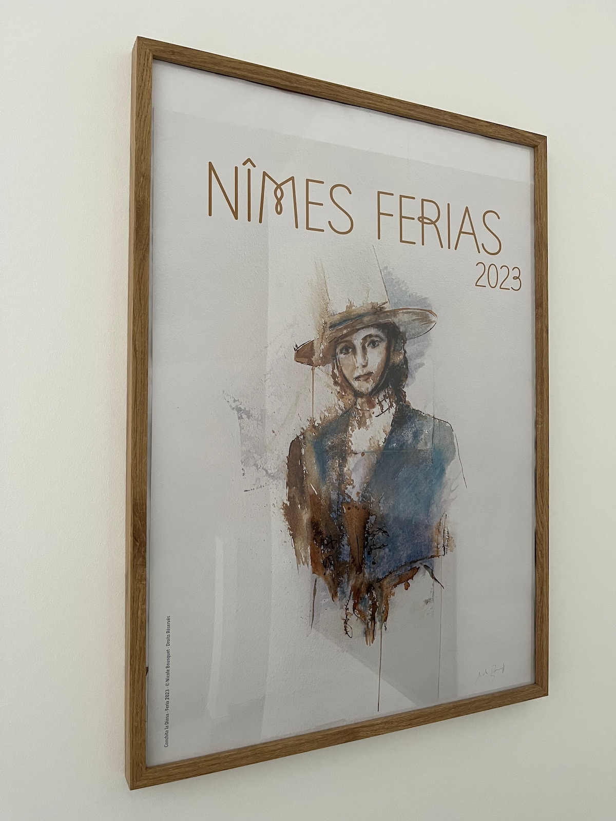 A framed art print featuring a watercolor depiction of a figure, is displayed on a light-colored wall. The artwork is titled 'NÎMES FÉRIAS 2023' in elegant gold lettering. The wooden frame complements the colors within the print.