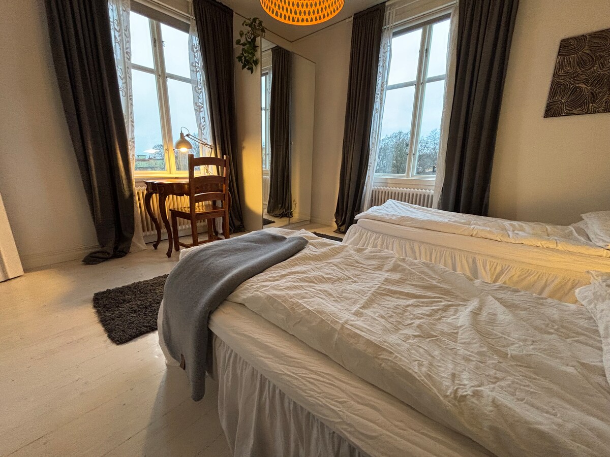 Calm bedroom with two single beds, soft lighting, and large windows framing treetop views. A small writing desk adds charm and function to this peaceful, light-filled space. A full-body mirror adds to the space.