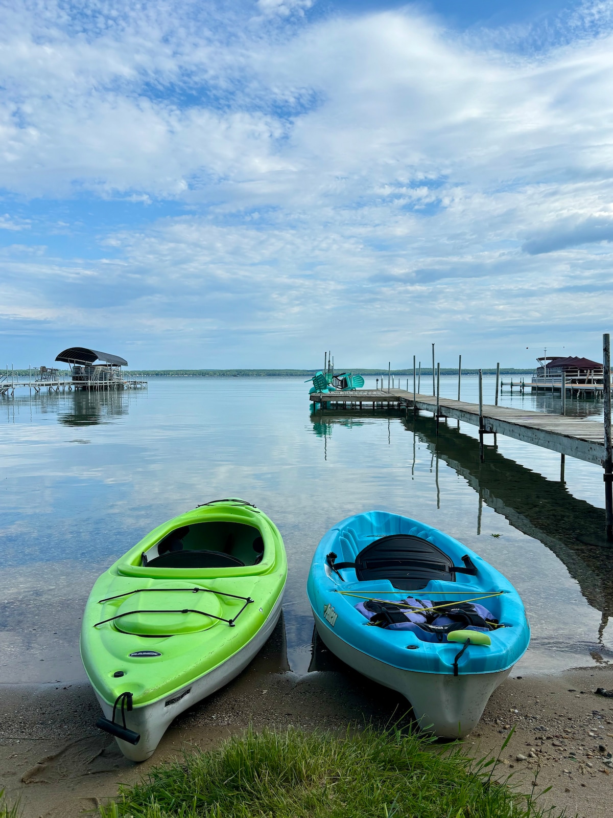 Two kayaks, one green and one blue, are positioned on a sandy shoreline with calm water reflecting the sky. A dock extends in the background alongside a boathouse, while low grass borders the scene, contributing to the serene lakeside environment.