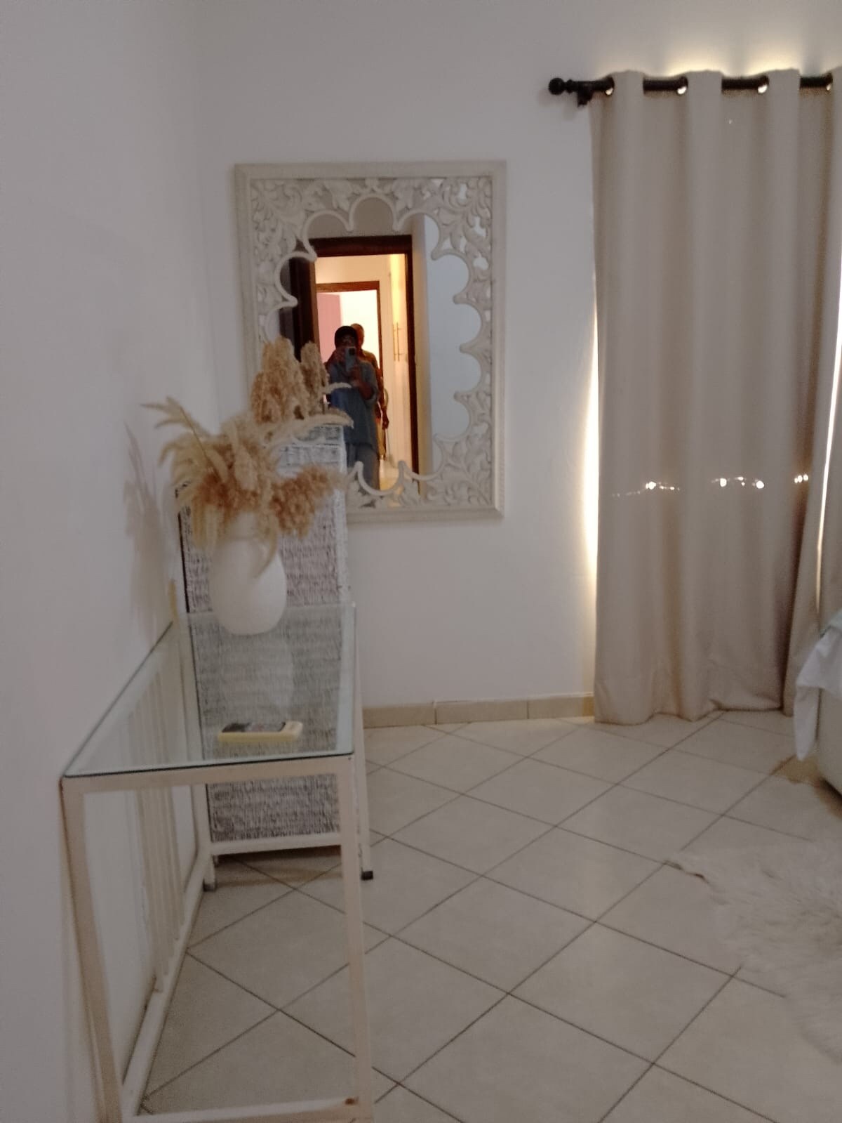 A bright entryway features a decorative mirror and a glass table with a vase of dried flowers. Soft curtains frame a doorway, allowing natural light to enter the space. The tile flooring adds a clean and open feel to the area.