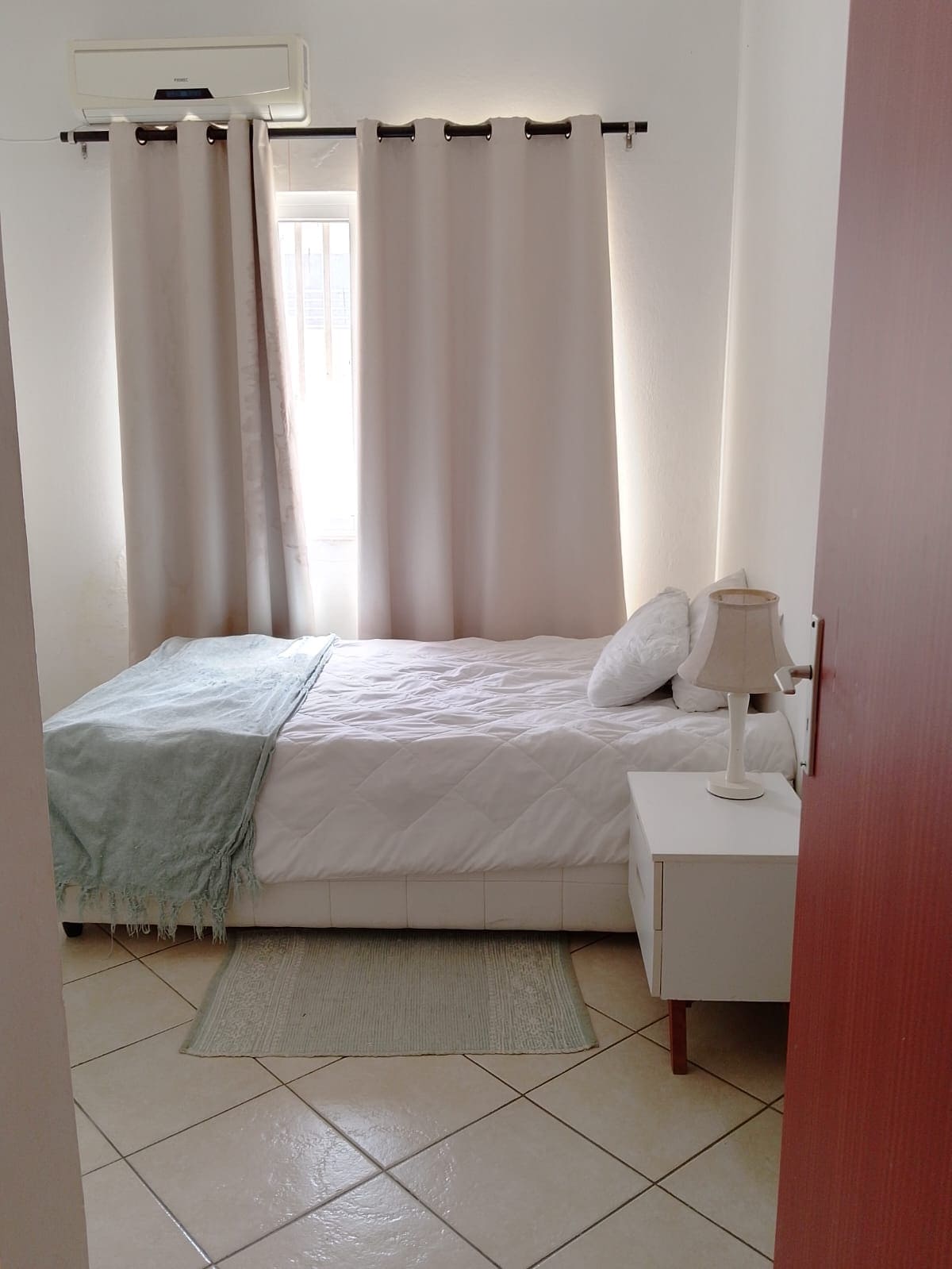 A serene sleeping area is accented by soft, neutral tones. A compact bed is dressed in a white comforter, accompanied by a light blue throw. A bedside table holds a simple lamp, and a window dressed with light curtains allows natural light to fill the space.