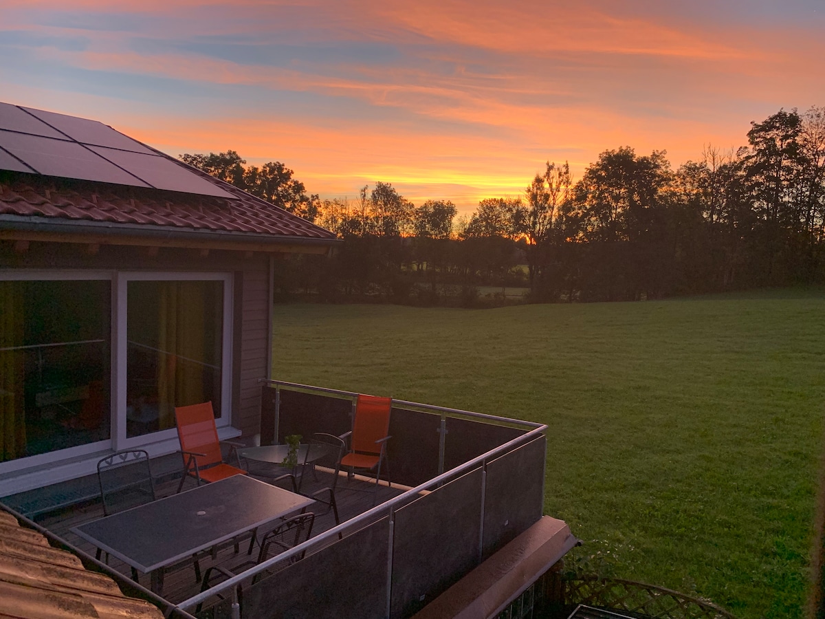 A terrace with two orange chairs and a dark table overlooks a sprawling green field. A sunset casts warm hues across the sky, with silhouettes of trees visible in the background. The house features large windows, allowing natural light to brighten the space.