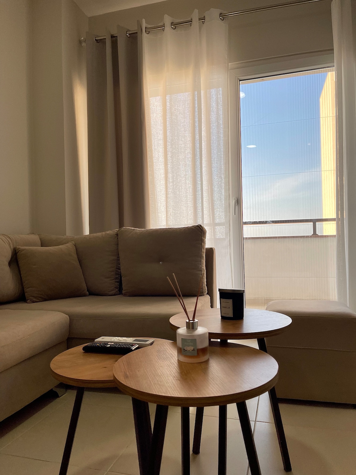 A comfortable seating area is highlighted by a soft, beige sofa and three round wooden side tables. Light filters through sheer curtains, illuminating the space. A small balcony is visible through the glass door, offering a glimpse of the outdoors.