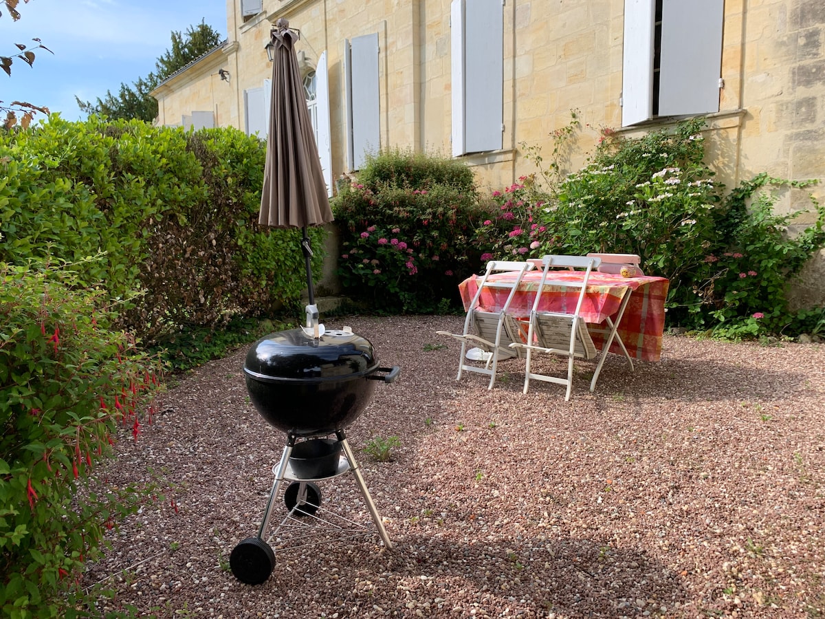 An outdoor space features a charcoal grill positioned beside a dining table draped in a vibrant tablecloth, surrounded by four white chairs. Lush greenery and blooming flowers create a natural border, while the facade of a stone building adds a rustic charm in the background.