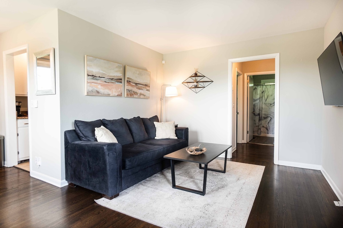 A spacious living area is characterized by a dark blue couch and a modern coffee table positioned on a light-colored area rug. Soft lighting is provided by a floor lamp, while artwork adorns the walls. Access to a hallway leading to the kitchen and bathroom is visible.