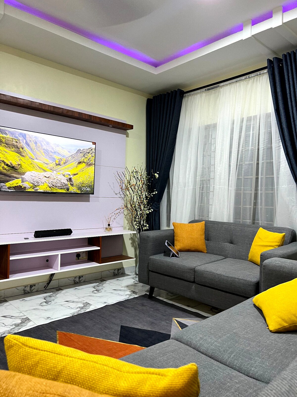 A cozy living area is furnished with a gray sectional sofa adorned with yellow cushions. A wall-mounted television displays scenic imagery, complemented by soft white drapes that filter natural light. The space features elegant flooring and a minimalistic decorative arrangement for a welcoming ambiance.