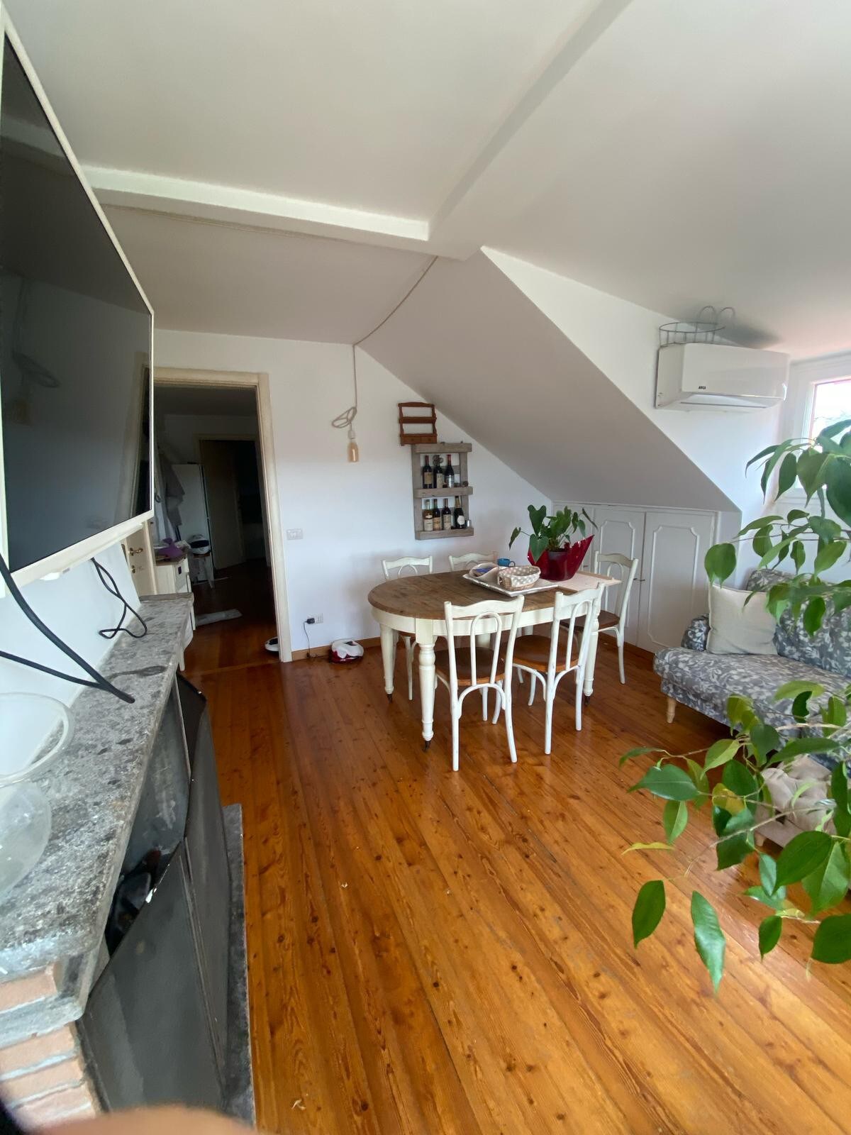 An open living space features a round dining table surrounded by six white chairs. A sizable television is mounted on the wall opposite a cozy seating area. Natural light is allowed in through a nearby window, illuminating the wooden flooring and enhancing the simplicity of the design.