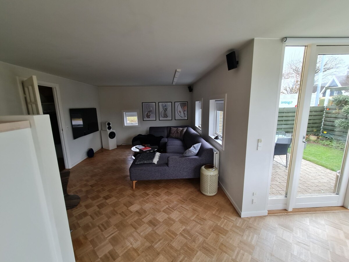 A cozy living area is presented with light-colored walls and parquet flooring. A spacious, dark gray sectional sofa provides ample seating, alongside a stylish floor lamp and decorative cushions. Windows allow natural light to fill the space, offering views of the garden outside.