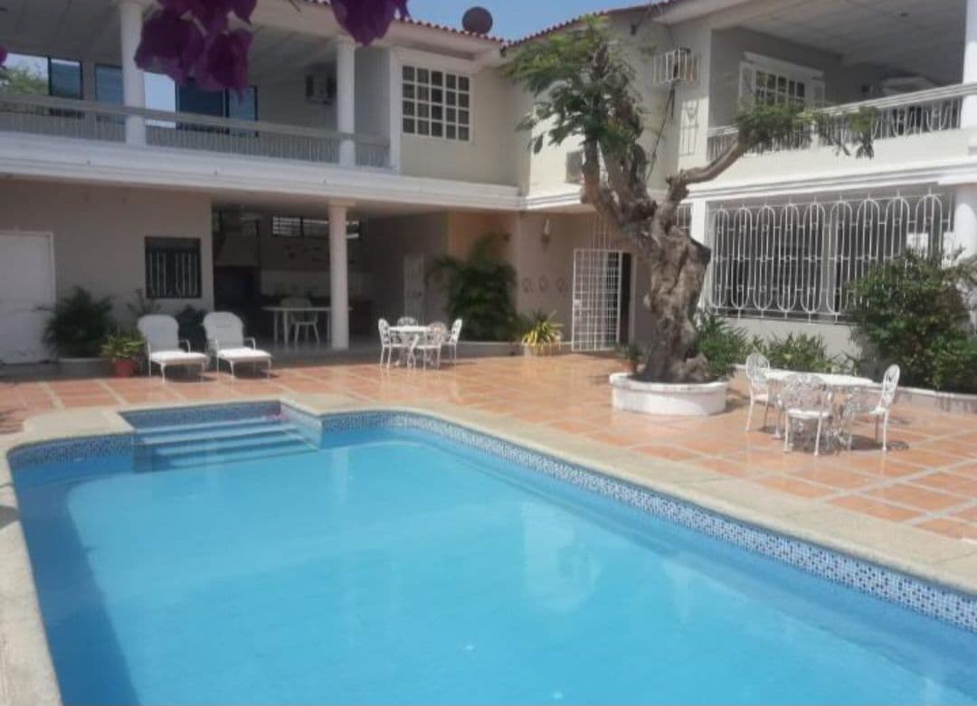 A well-maintained pool occupies the center of a tiled patio, surrounded by lounge chairs. In the background, a spacious terrace with tables and chairs can be seen, set against a backdrop of lush greenery and a mature tree.